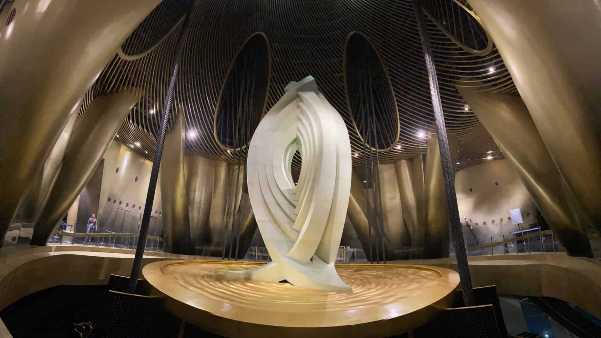 Interior do Shanghai Tower, mostrando o amortecedor de massa sintonizada. A estrutura metálica curvada e dourada envolve a sala, com cabos de aço conectados ao amortecedor para estabilizar o edifício. No centro da imagem, há uma escultura moderna, iluminada por luzes no teto, e uma pessoa pequena ao fundo para referência de escala.