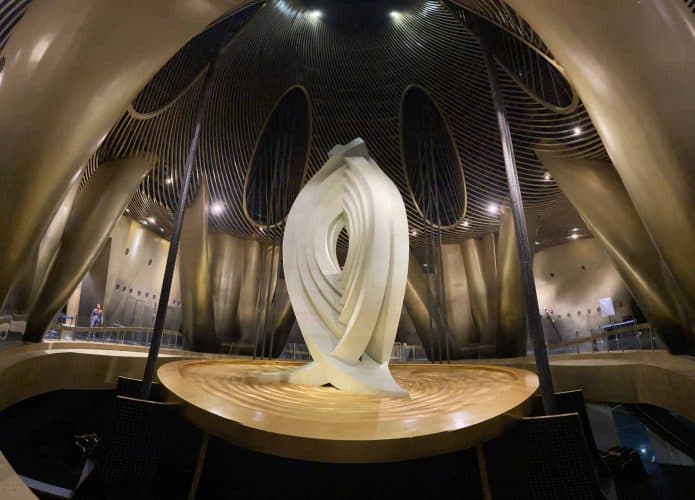 Interior do Shanghai Tower, mostrando o amortecedor de massa sintonizada. A estrutura metálica curvada e dourada envolve a sala, com cabos de aço conectados ao amortecedor para estabilizar o edifício. No centro da imagem, há uma escultura moderna, iluminada por luzes no teto, e uma pessoa pequena ao fundo para referência de escala.