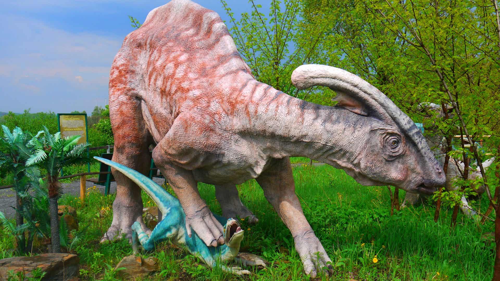 Escultura de dinossauro Parasaurolophus pisando em dinossauro menor enquanto anda em floresta