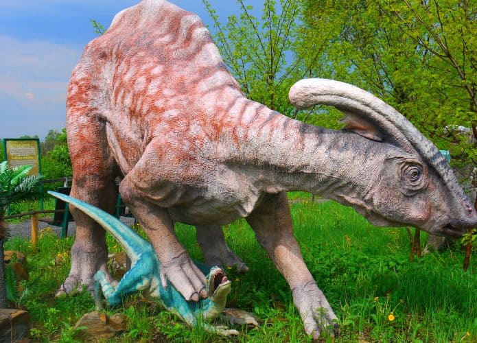 Escultura de dinossauro Parasaurolophus pisando em dinossauro menor enquanto anda em floresta