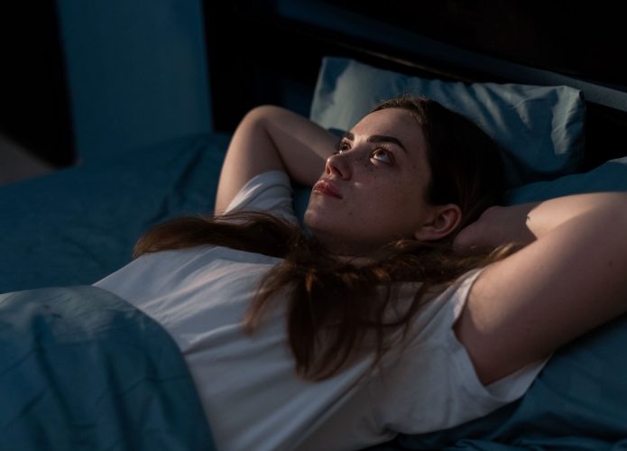 Mulher deitada na cama olhando para o teto com os braços cruzados atrás da cabeça
