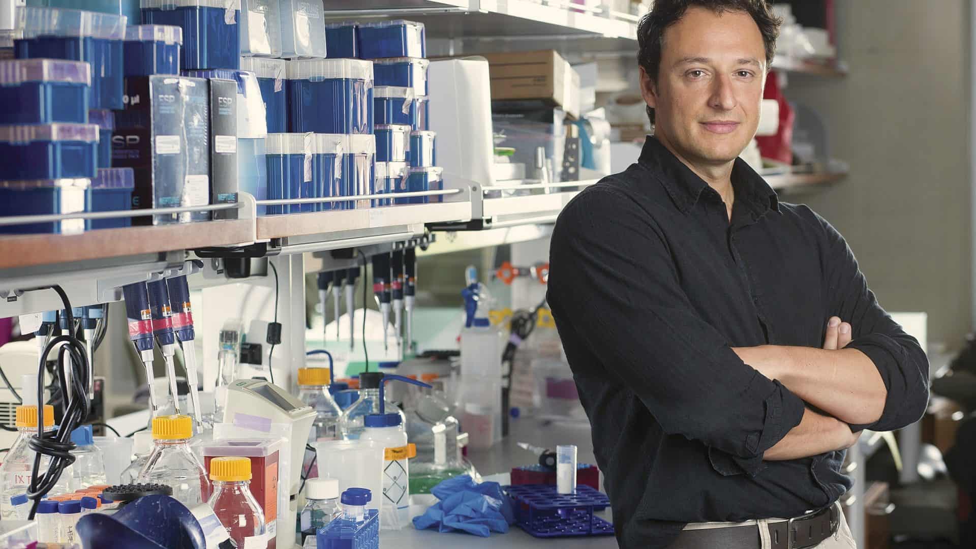 Biólogo, com os braços cruzados, escorado em uma bancada em seu laboratório
