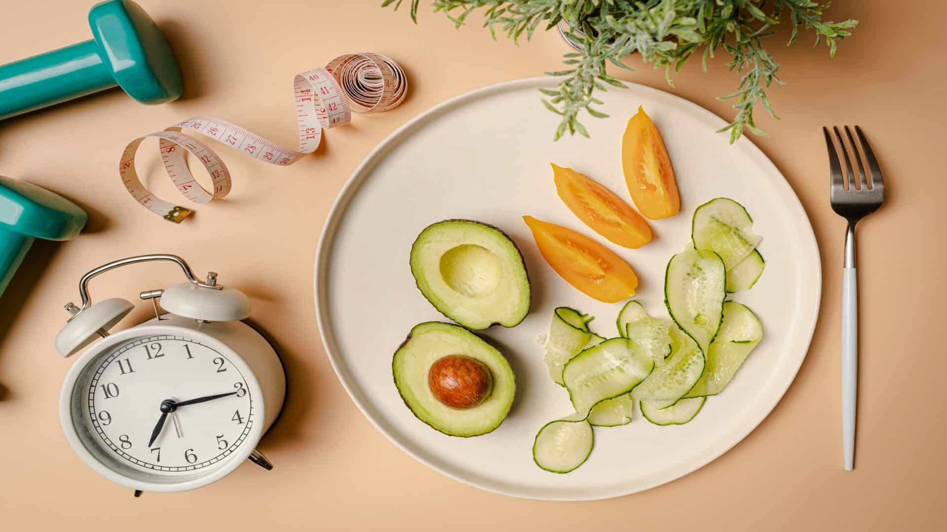 Composição criativa em flat lay com prato, despertador, faca, garfo, haltere e fita métrica sobre fundo rosa. Fasting intermitente, cetogênica, conceito de dieta. vista de cima, espaço para texto.
