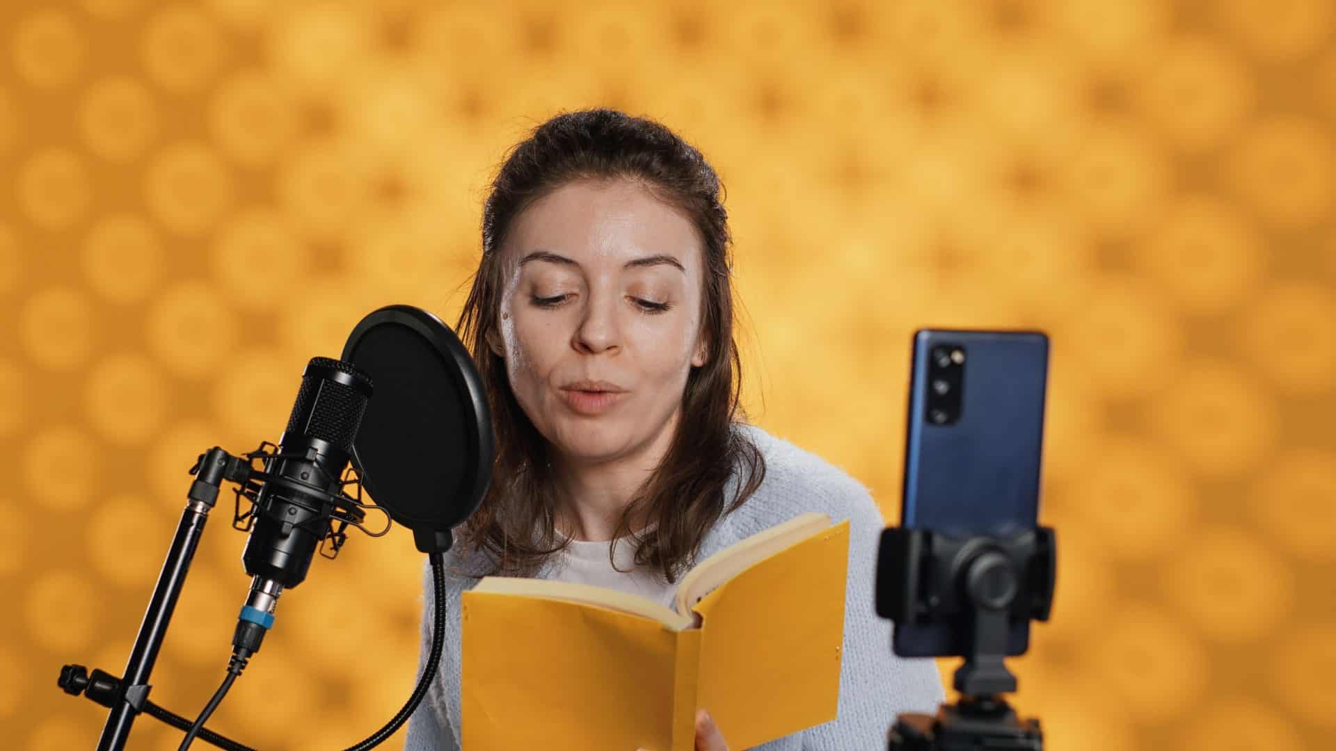 Narrador olhando para a câmera do telefone, filmando a si mesmo lendo um livro para crianças, criando audiobook, fundo de estúdio. Criador de conteúdo fazendo vlog enquanto produz gravação digital de romance infantil, câmera A