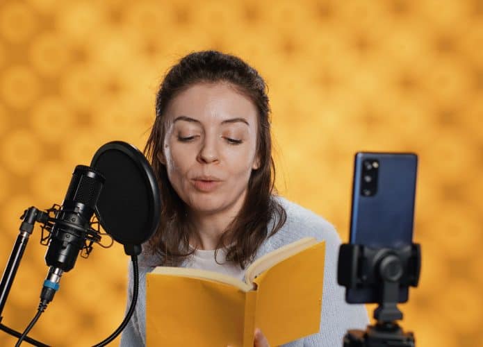 Narrador olhando para a câmera do telefone, filmando a si mesmo lendo um livro para crianças, criando audiobook, fundo de estúdio. Criador de conteúdo fazendo vlog enquanto produz gravação digital de romance infantil, câmera A