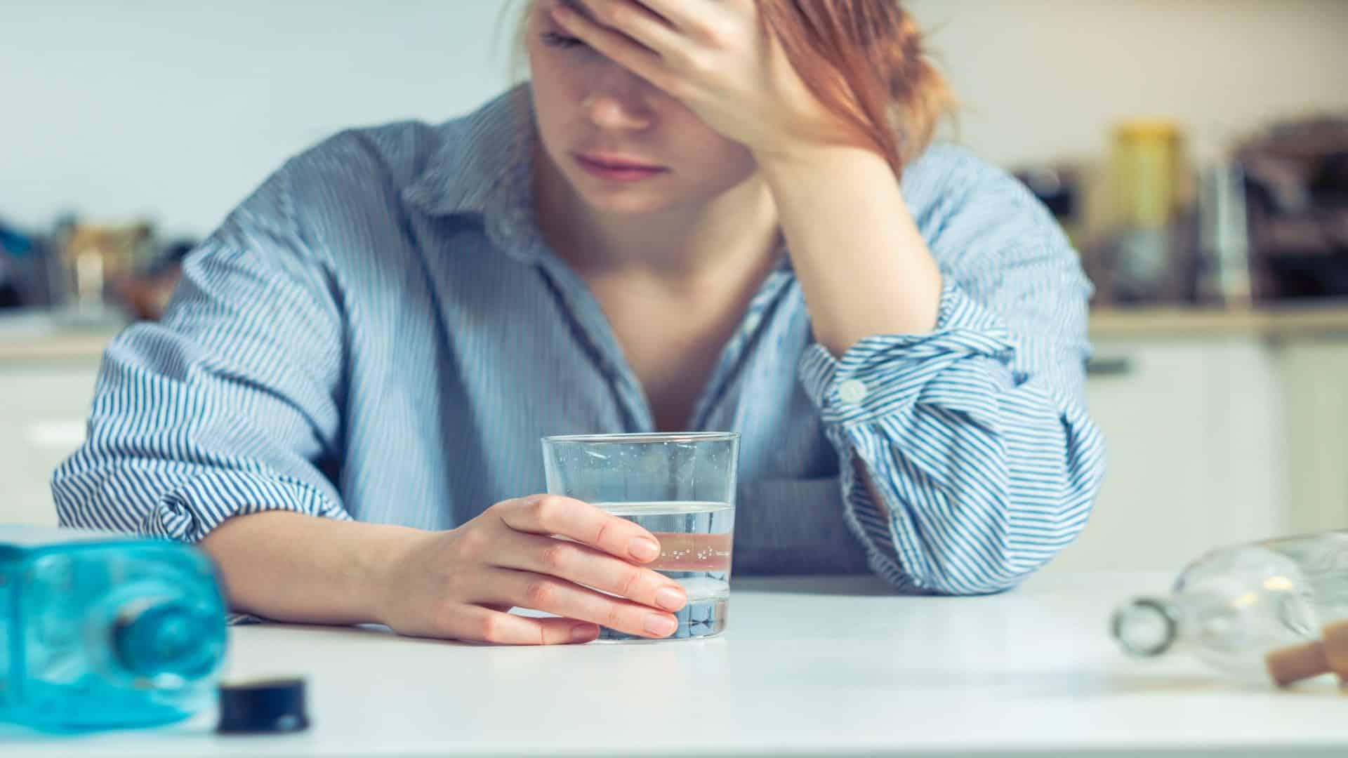 Uma mulher sofre de ressaca: ela se senta à mesa, segura a cabeça, há um copo de água e um pacote de comprimidos perto, uma expressão de dor e fadiga em seu rosto.