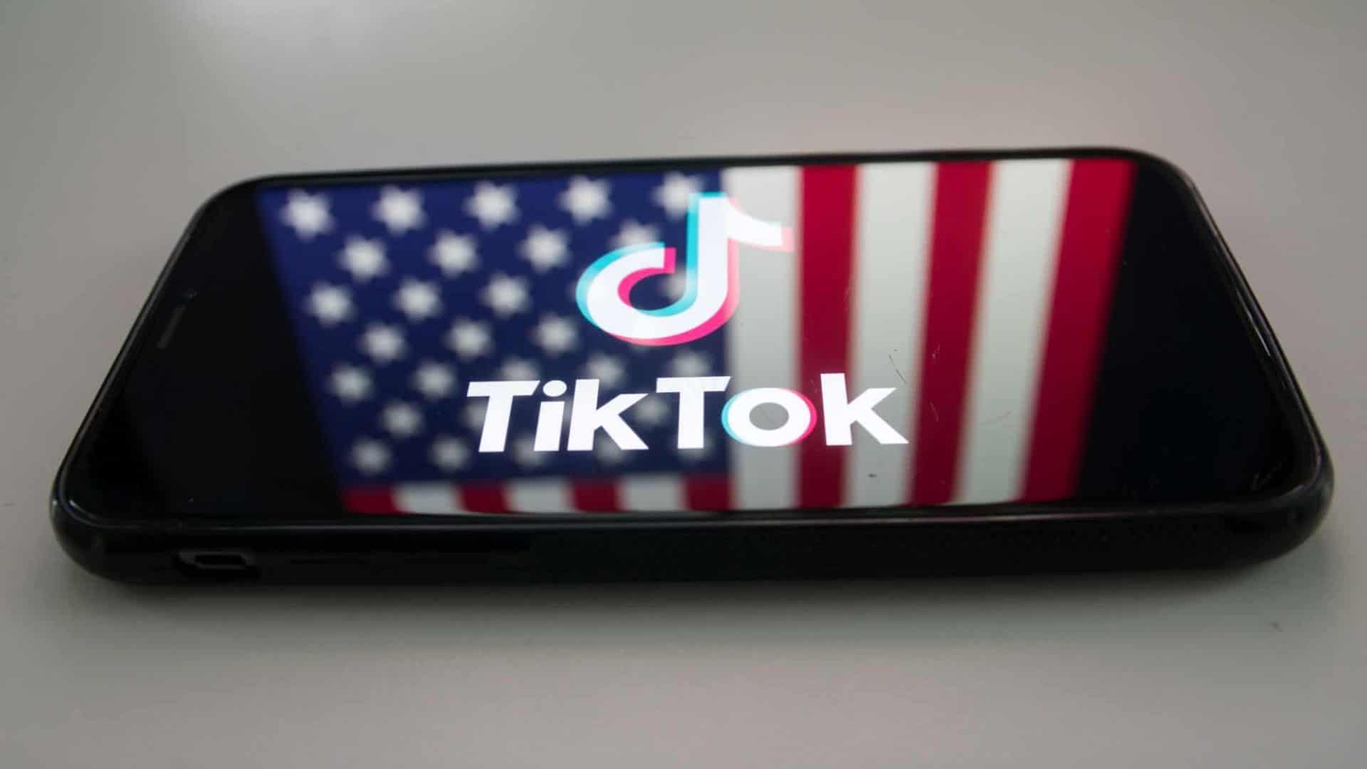 Logo do TikTok refletido em uma tela de smartphone que possui, de fundo, a bandeira dos Estados Unidos