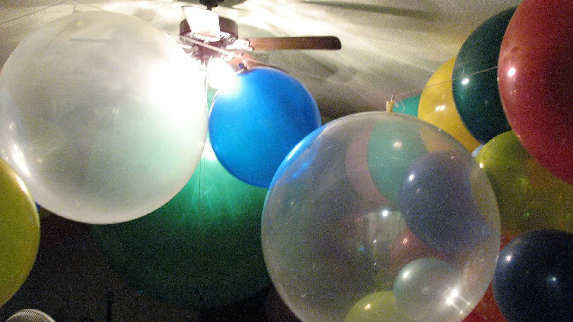 Um quarto repleto de balões de cores variadas, incluindo balões grandes e transparentes, além de balões vermelhos, azuis, verdes e amarelos. O teto é baixo, e no centro há um ventilador de teto iluminado.