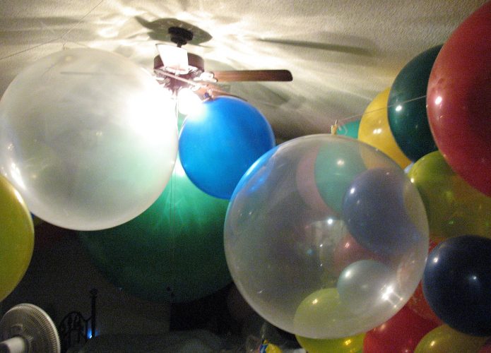 Um quarto repleto de balões de cores variadas, incluindo balões grandes e transparentes, além de balões vermelhos, azuis, verdes e amarelos. O teto é baixo, e no centro há um ventilador de teto iluminado.