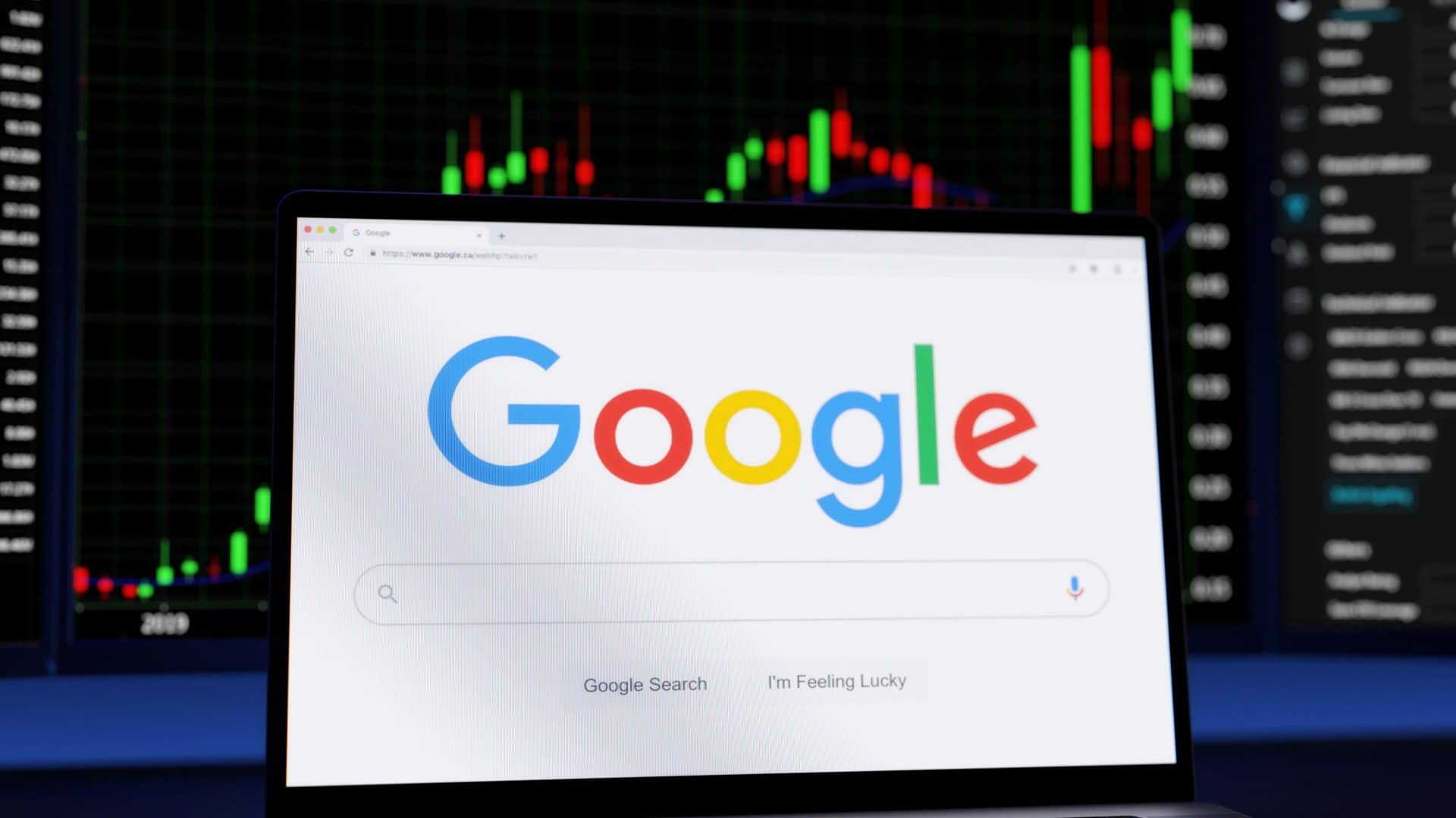 Página inicial do Google aberta em notebook colocado na frente de monitor com linhas vermelhas e verdes de ações financeiras