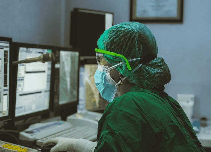 imagem mostra uma mulher paramentada com EPIs registrando um prontuário médico em um computador num hospital