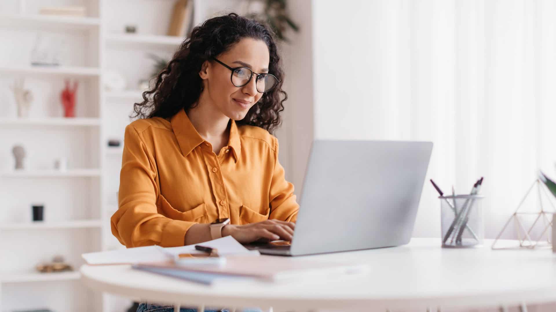 imagem mostra uma mulher sentada a uma mesa, mexendo em seu notebook