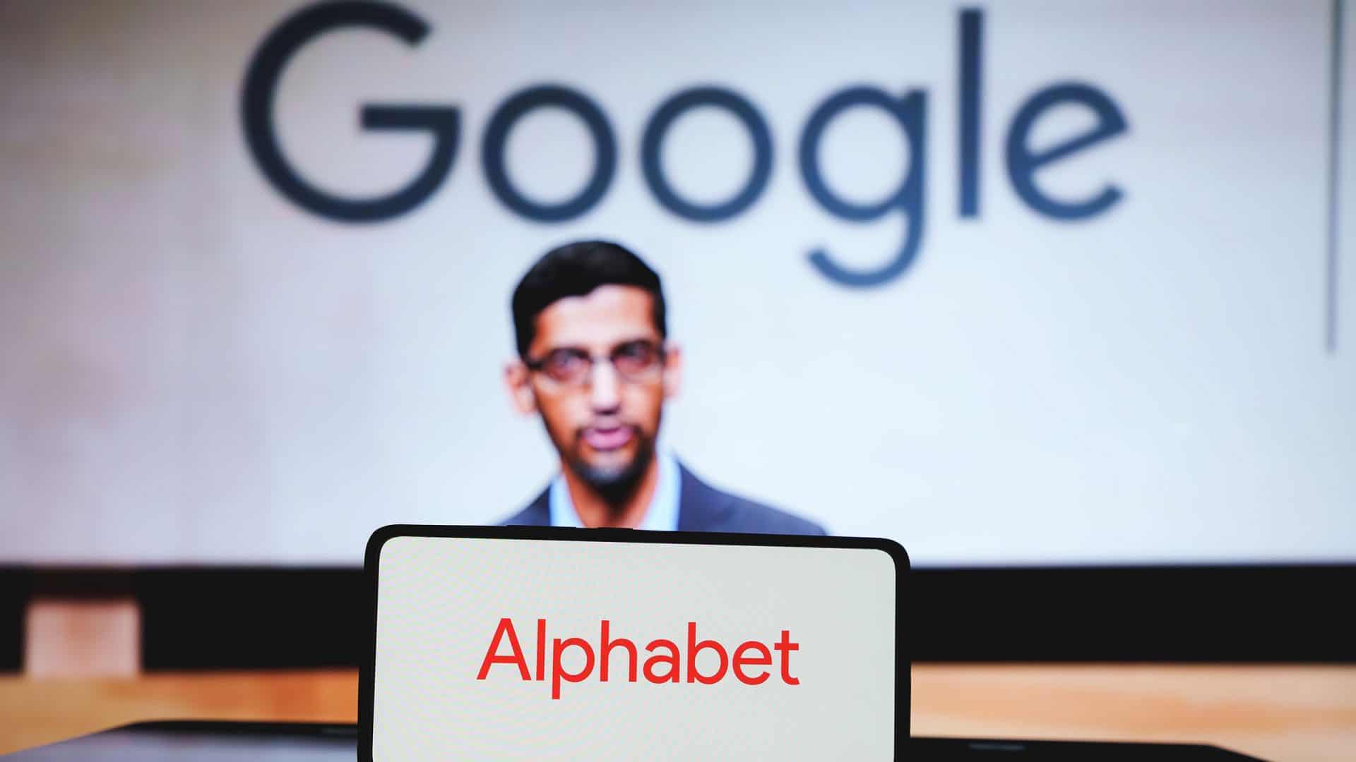 Celular com logomarca da Alphabet na tela colocado na frente de monitor exibindo foto do CEO do Google, Sundar Pichai, na frente de letreiro da empresa