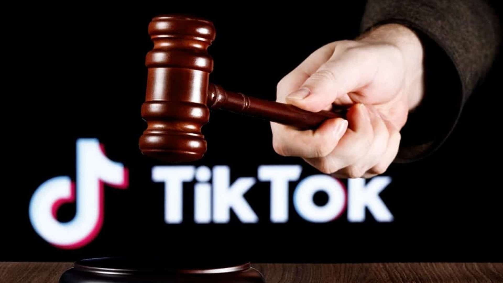 À frente, um martelo de juiz segurado por uma mão; abaixo, uma mesa; atrás, desfocado, o logotipo do TikTok