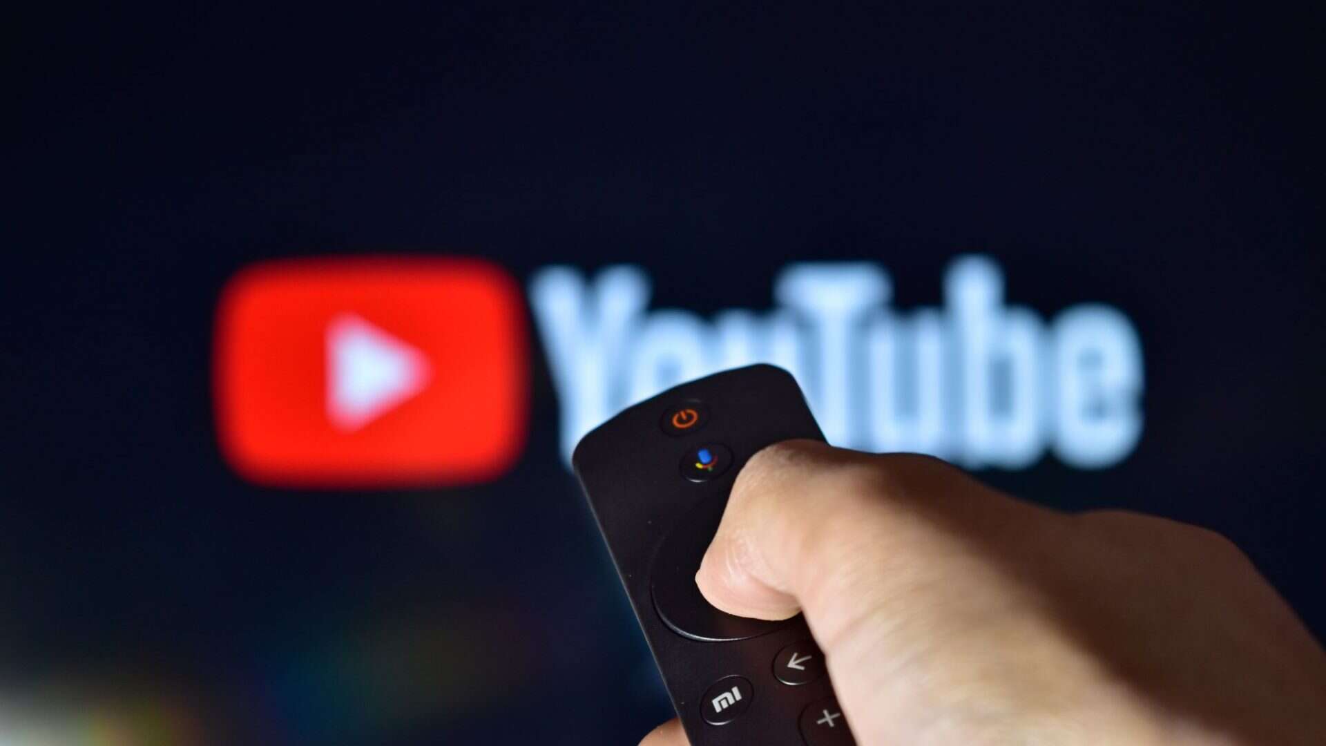 Logo do YouTube ao fundo e desfocado, com uma mão segurando um controle remoto à frente