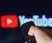 Logo do YouTube ao fundo e desfocado, com uma mão segurando um controle remoto à frente