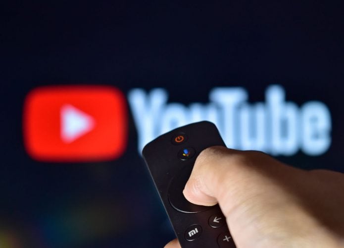 Logo do YouTube ao fundo e desfocado, com uma mão segurando um controle remoto à frente