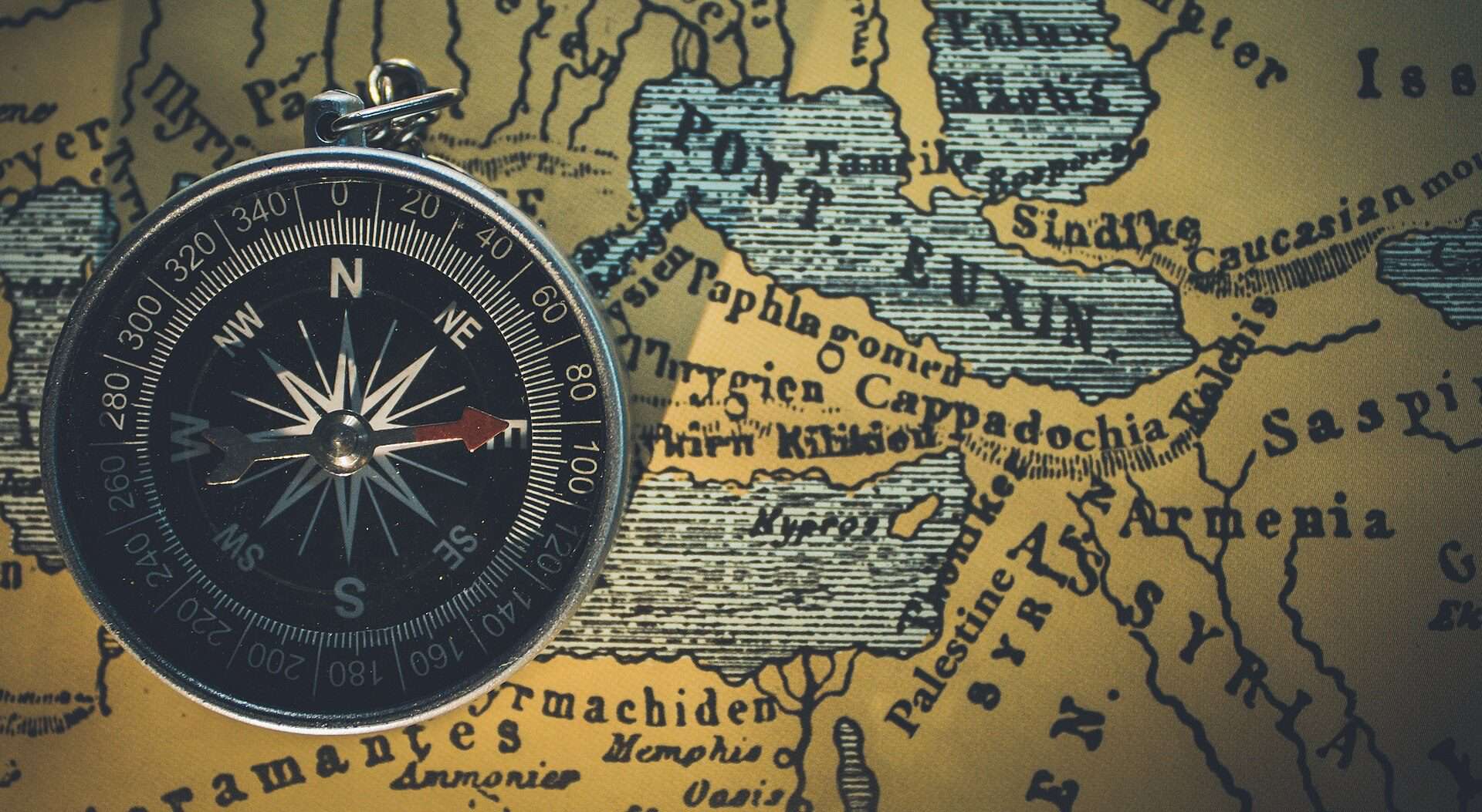 Uma bússola metálica em close-up, com ponteiros apontando para o norte e o sul, colocada sobre um mapa histórico que destaca regiões antigas como Armênia e Síria. Crédito: Himesh Kumar Behera, thehopefultraveller, CC0, via Wikimedia Commons.