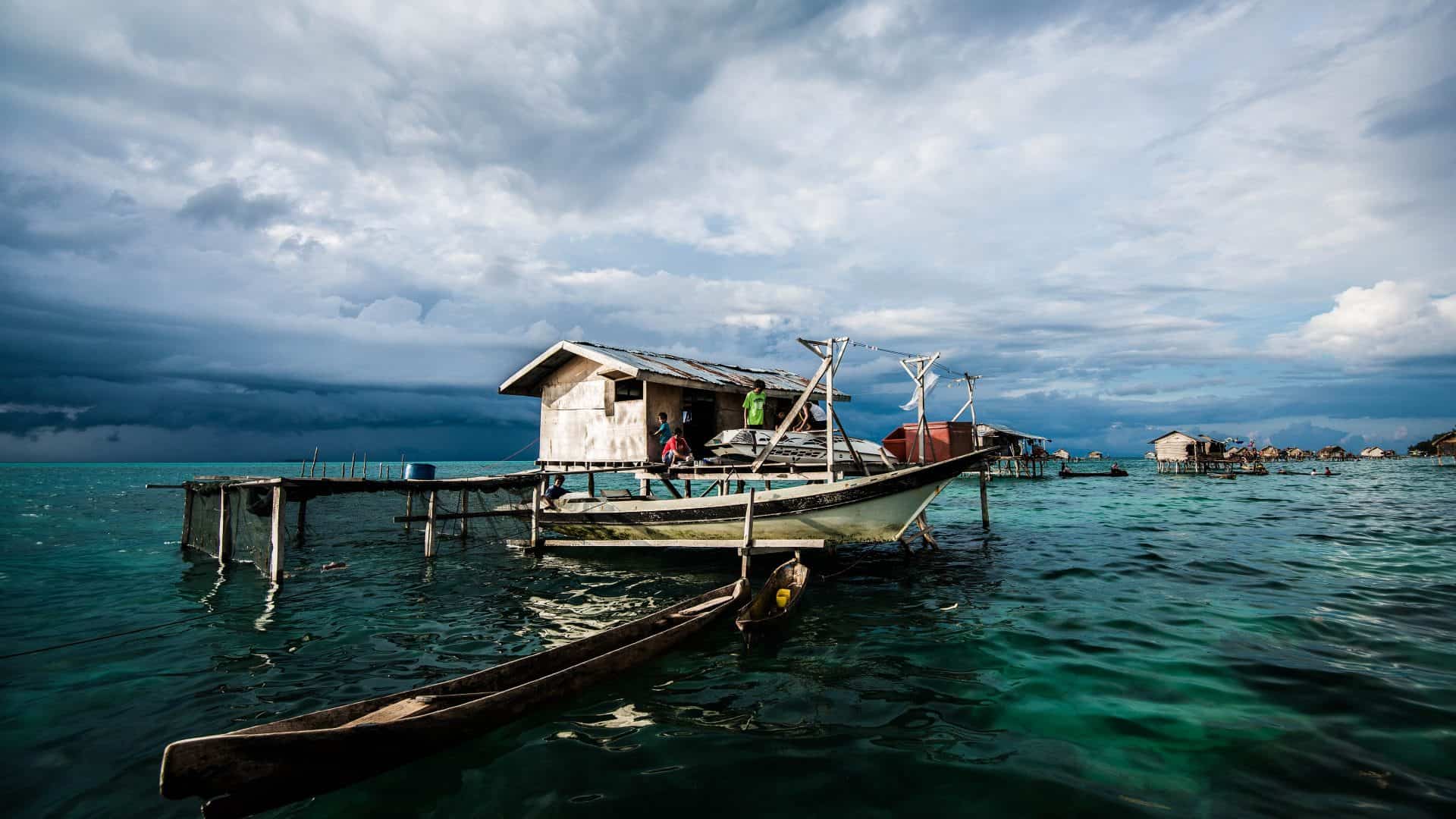 Casas Bajau construídas sobre palafitas no meio do oceano, com barcos de madeira ancorados e pessoas realizando atividades do cotidiano. O céu ao fundo está nublado, criando uma atmosfera dramática sobre as águas cristalinas.