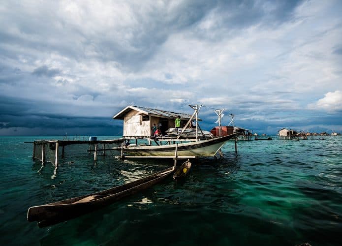 Casas Bajau construídas sobre palafitas no meio do oceano, com barcos de madeira ancorados e pessoas realizando atividades do cotidiano. O céu ao fundo está nublado, criando uma atmosfera dramática sobre as águas cristalinas.