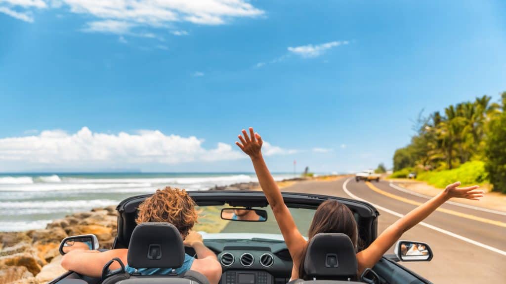 Viagem de carro férias felizes casal dirigindo um carro conversível nas férias de verão no Havai. Mulher com os braços para cima se divertindo, jovem homem ao volante.