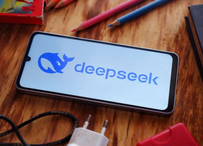 Celular com logotipo do DeepSeek na tela colocado sobre mesa de madeira rodeado de materiais de escritório como lápis e carregador