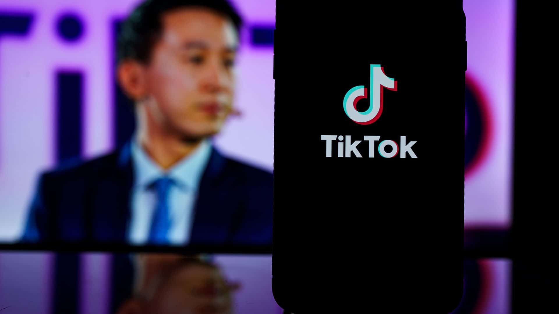 Ao fundo, foto desfocada de Shou Chew; à frente logotipo do TikTok em um smartphone, que está em pé em cima de uma mesa
