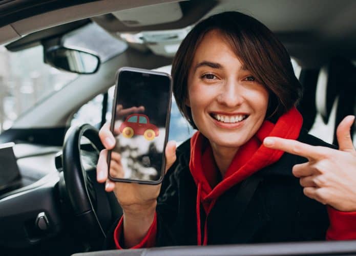 Mulher dentro de um carro, no banco do motorista, sorrindo e segurando o celular com um ícone de carro na tela, representando o uso de apps de transporte