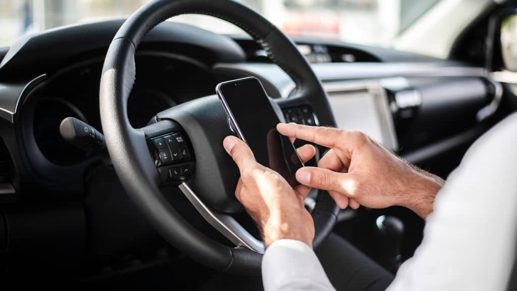 Motorista imerso no trânsito, com as mãos no volante e mexendo no celular, enquanto o fundo desfocado destaca o cenário urbano