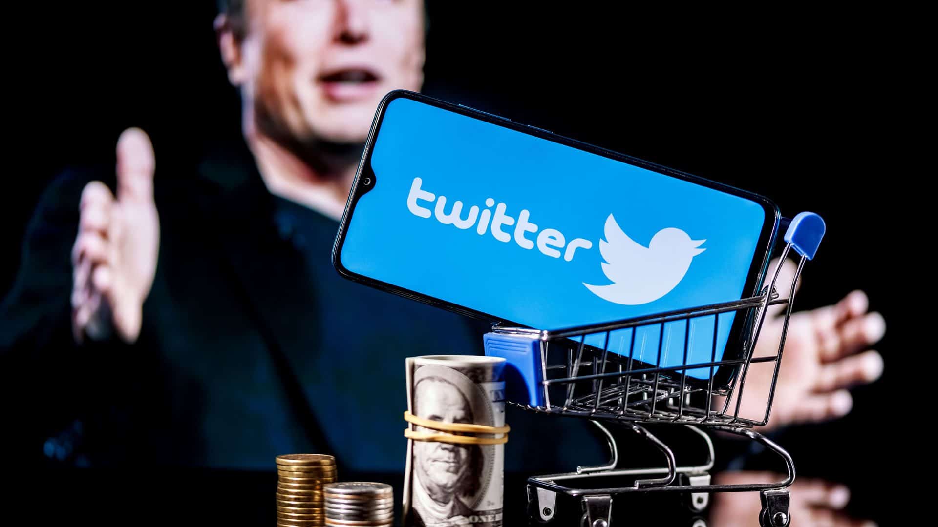 Ao fundo, Elon Musk falando; mais à frente, logo do antigo Twitter exibido em um smartphone; em primeiro plano, um carrinho de compras, moedas e notas de dinheiro