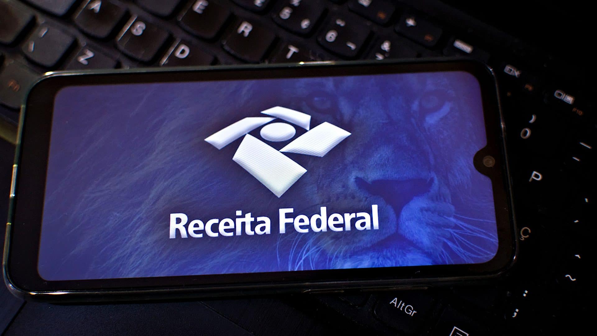 Logo da Receita Federal exibido em um smartphone que está em cima de um teclado de computador