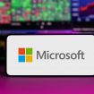 Logo da Microsoft em um smartphone; atrás, telas com gráficos com ações
