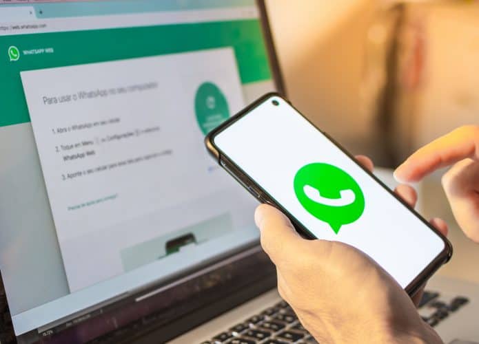 Mãos segurando um dispositivo de telefone celular inteligente com o logotipo do aplicativo da empresa WhatsApp na tela e um notebook com o aplicativo da Web WhatsApp no ​​navegador da Internet