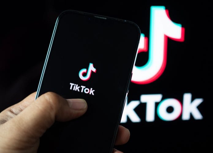 Na imagem, uma mão segura um celular cuja tela apresenta o logo do TikTok