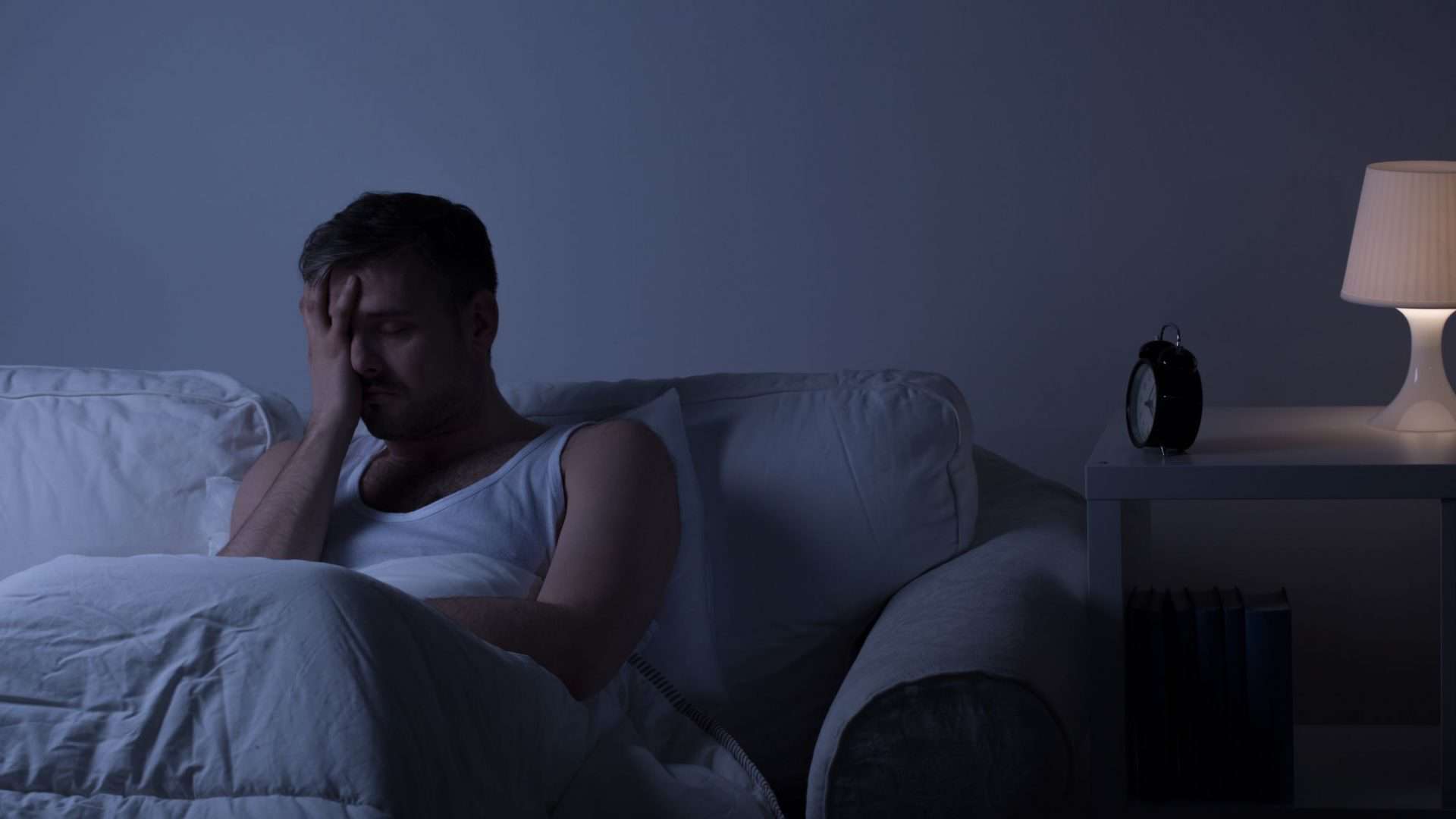 Homem sentado em uma cama, à noite, com a mão no rosto