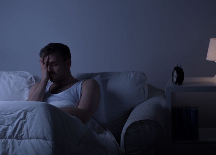 Homem sentado em uma cama, à noite, com a mão no rosto