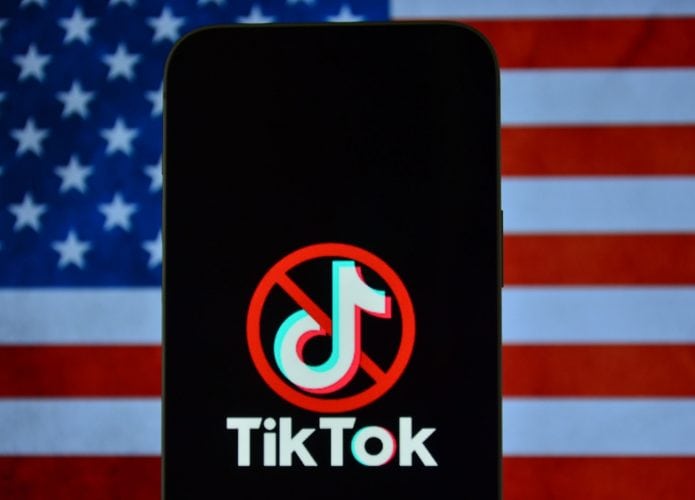 Logo do TikTok com um ícone de proibição por cima em um smartphone; atrás, a bandeira dos Estados Unidos