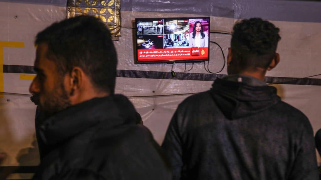 Foto com aparelho de TV ao fundo e um homem de costas assistindo notícia sobre cessar fogo entre Israel e Hamas, e outro homem passa ndo.