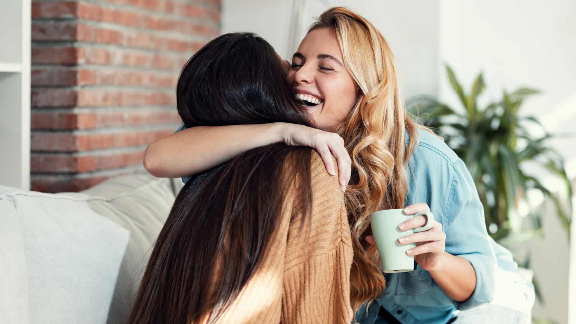 Imagem de duas mulheres adoráveis sorrindo e se abraçando enquanto conversam sentadas no sofá na sala de estar em casa.