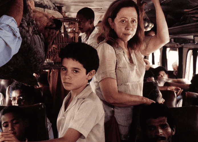 Uma mulher e um menino estão de pé em um ônibus lotado. Ela segura no suporte enquanto ele olha sério para a câmera. Passageiros e bagagens preenchem o espaço