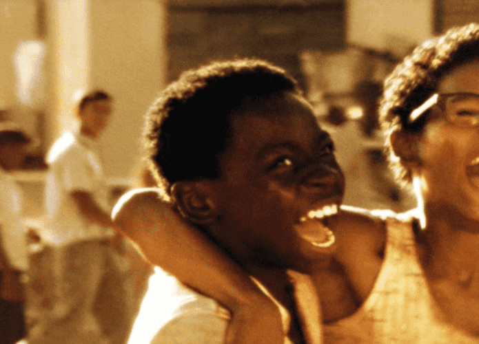Dois jovens negros riem com entusiasmo enquanto um abraça o outro pelo ombro. A cena, em tons quentes, transmite alegria e energia, com figuras desfocadas ao fundo em um ambiente de periferia.