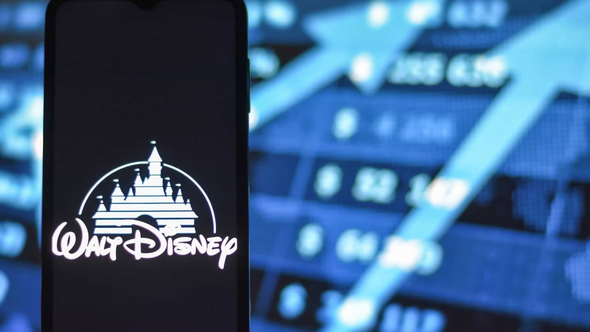 Logo da Walt Disney em um smartphone; ao fundo, gráficos com ações