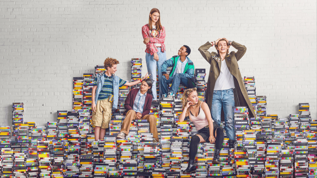 Imagem promocional da série 'Everything Sucks!'. Seis adolescentes posam sobre uma pilha de fitas VHS, formando uma escadaria. O fundo é um muro branco. Cada personagem tem um estilo distinto, refletindo a estética dos anos 90.