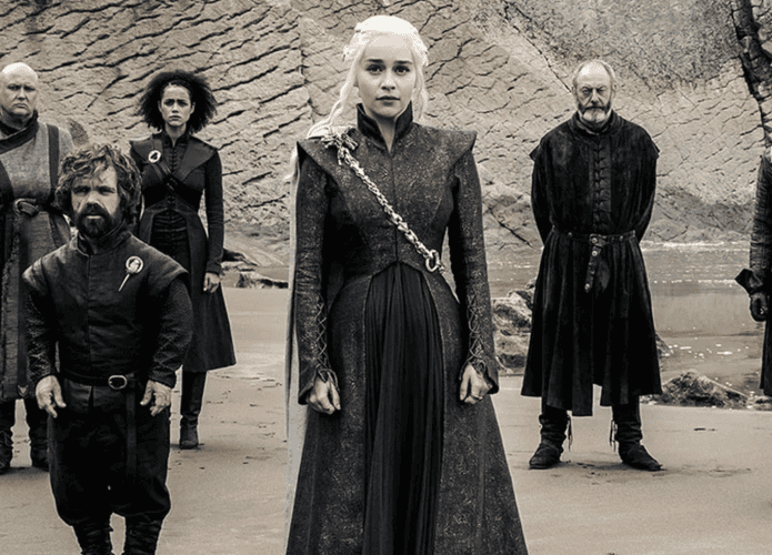 Cena de Game of Thrones com Daenerys Targaryen em primeiro plano, cercada por Tyrion Lannister, Jon Snow, Varys, Missandei e Sor Davos em uma praia com rochas ao fundo