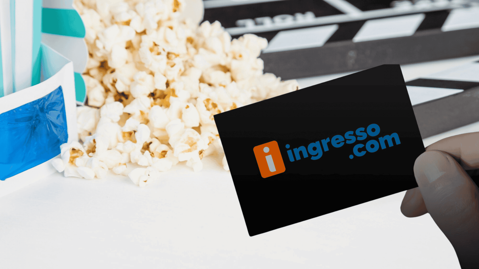 Cartão preto com o logotipo da Ingresso.com sendo segurado por uma mão. Ao fundo, há pipoca espalhada sobre uma superfície branca e uma claquete de cinema