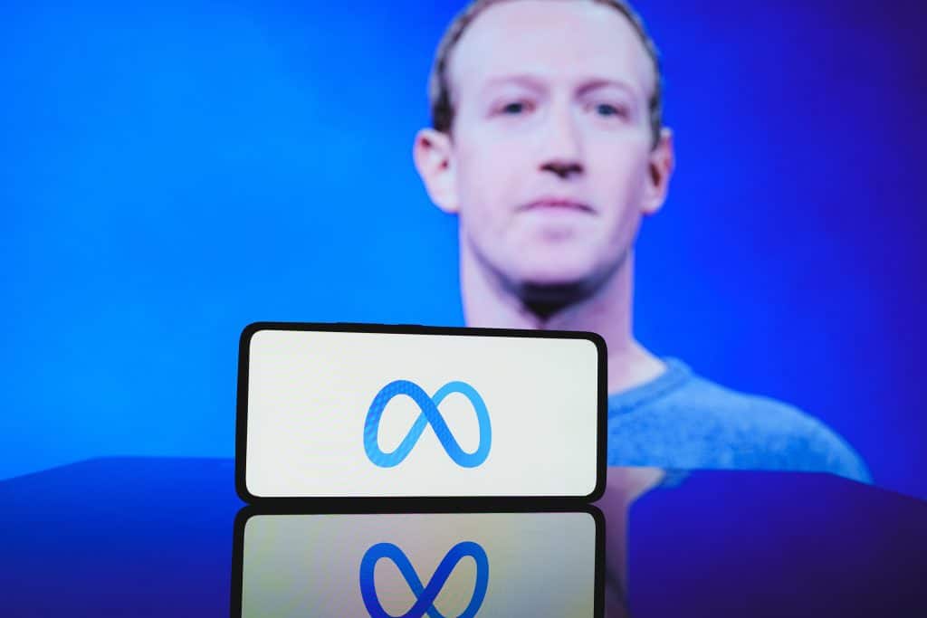 Ao fundo, foto desfocada de Mark Zuckerberg; à frente, logo da Meta em um smartphone