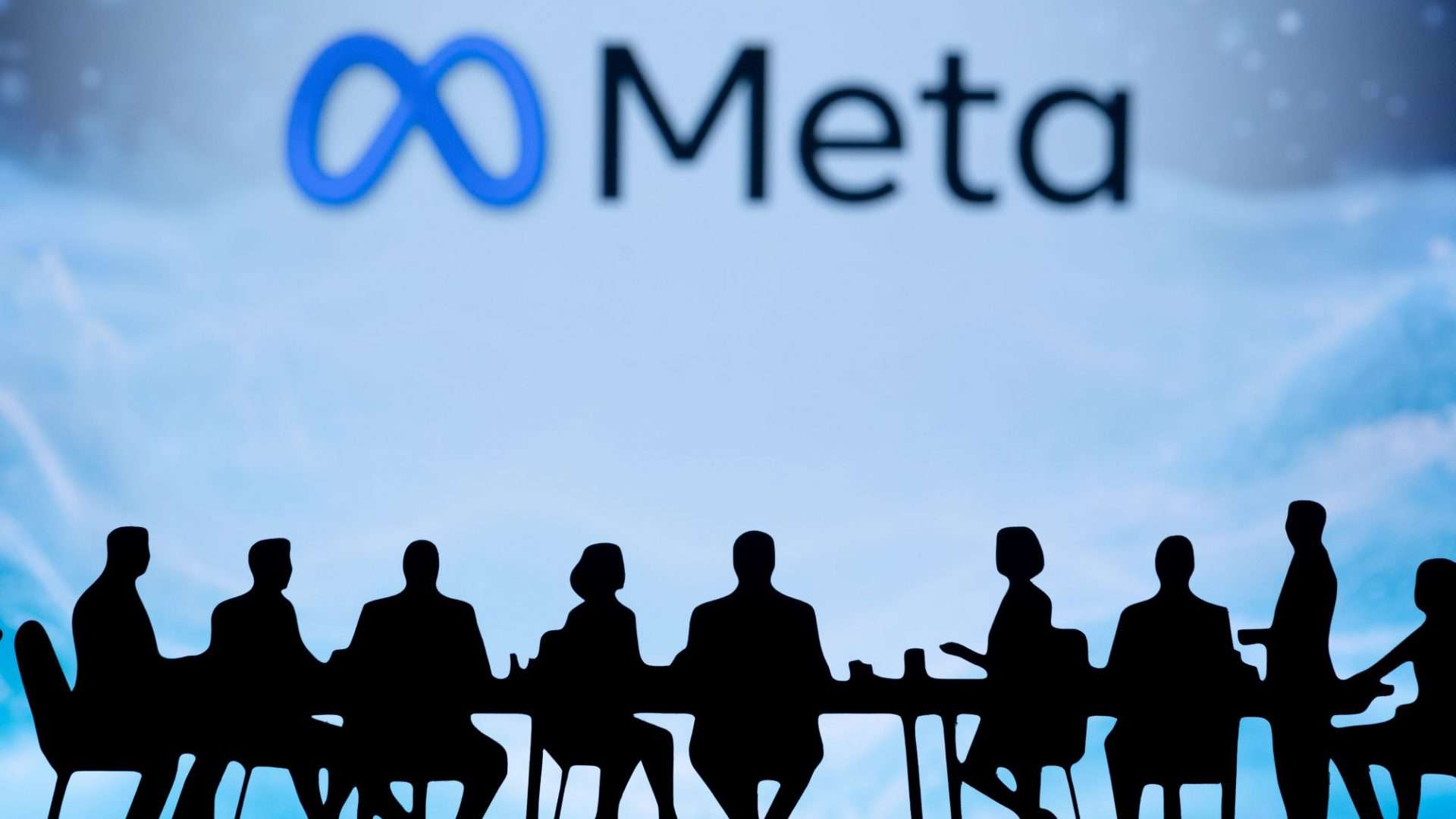 Ao fundo, em um telão e meio desfocado, logo da Meta; à frente, sombras de várias pessoas sentadas em uma mesa