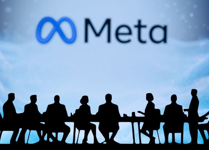 Ao fundo, em um telão e meio desfocado, logo da Meta; à frente, sombras de várias pessoas sentadas em uma mesa