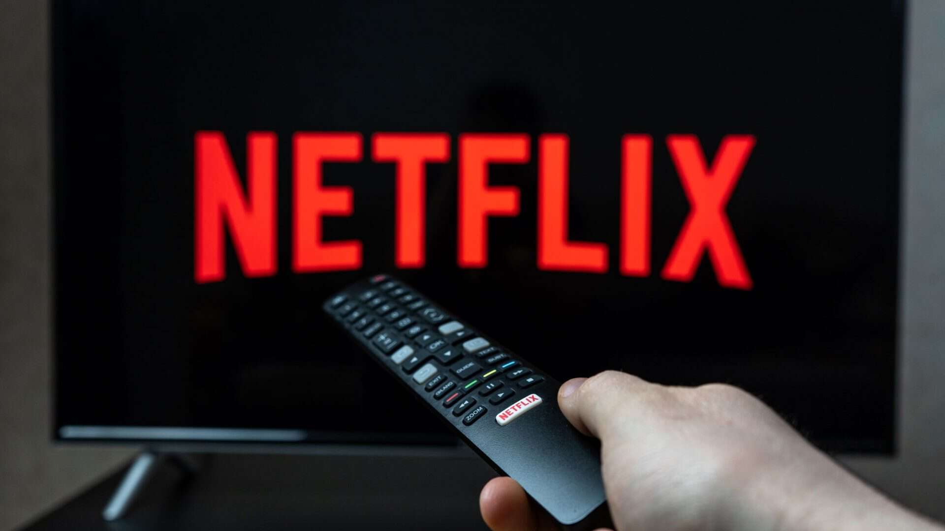 Logo da Netflix em uma TV; à frente, uma pessoa segurando um controle remoto