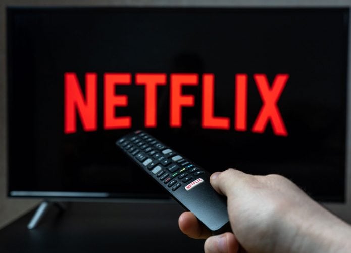 Logo da Netflix em uma TV; à frente, uma pessoa segurando um controle remoto